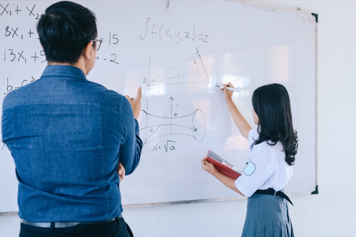 A student writing on a whiteboard to work through a problem, with her teacher alongside her.